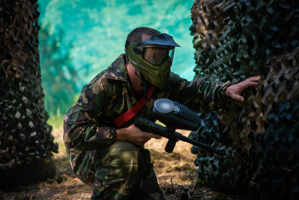 Man in camouflage suit and a mask hides behind camouflage wall with paintball gun